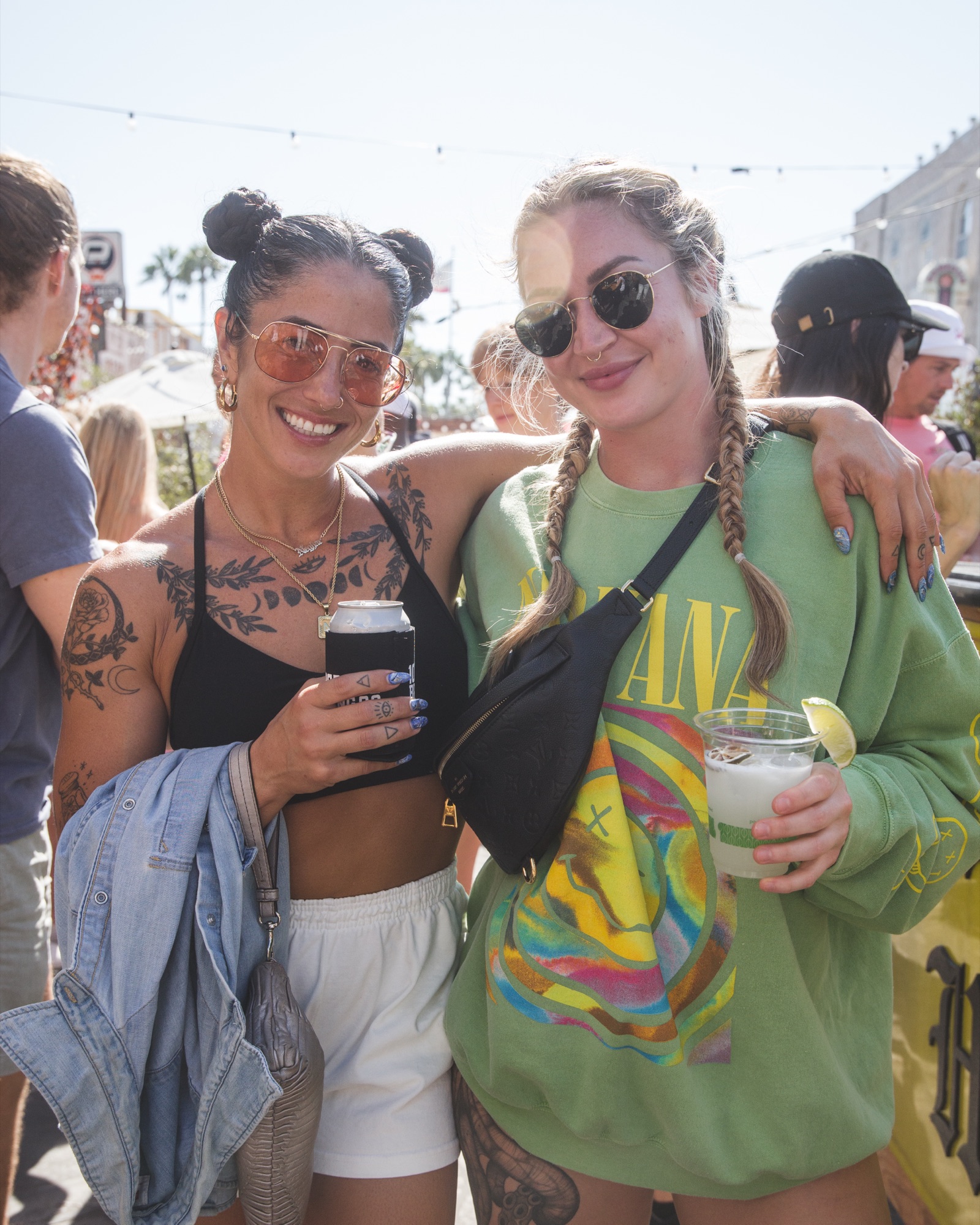 Two friends grinning at the beer garden, drinks in hand, sun overhead, the boardwalk behind them.