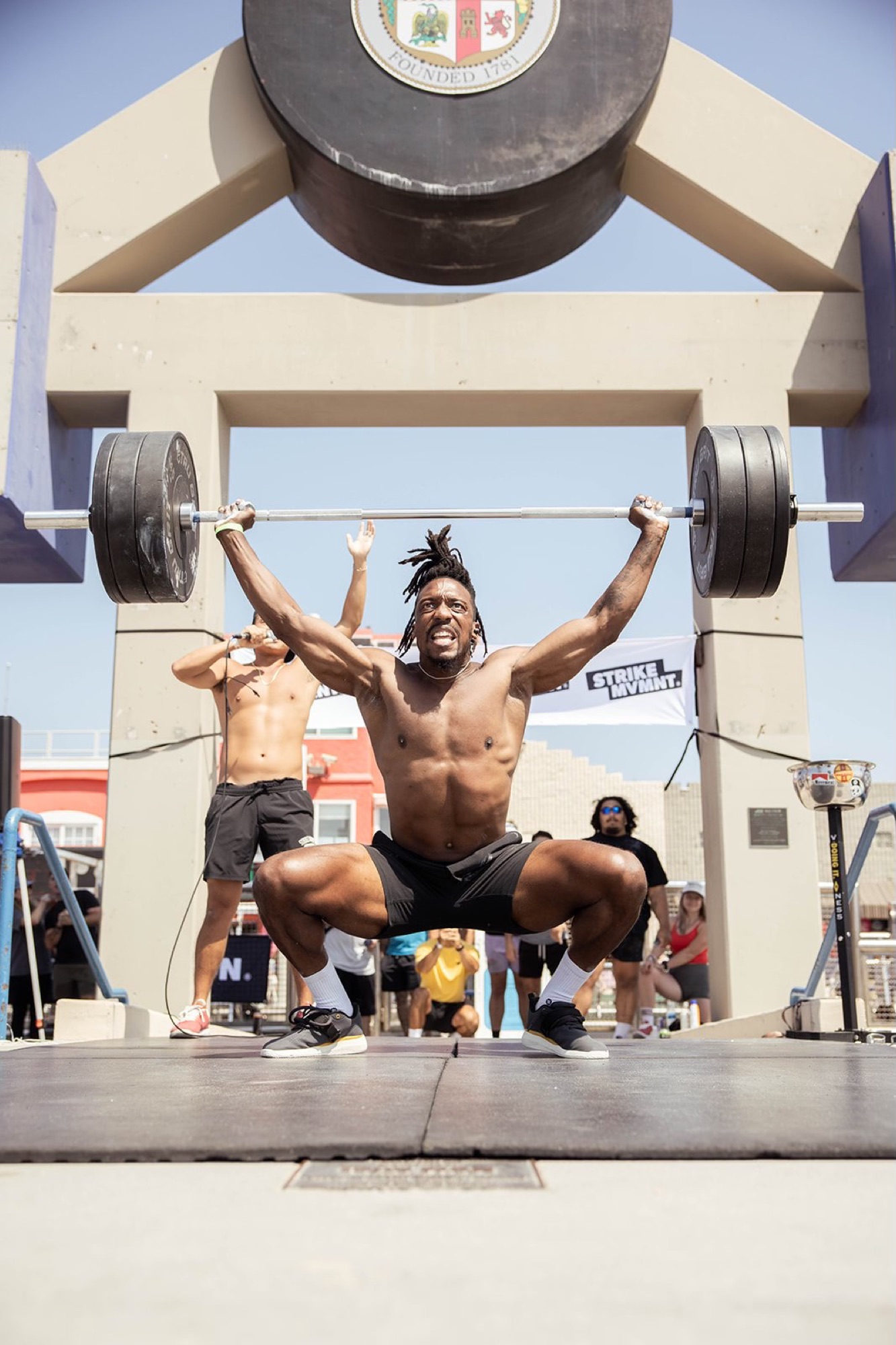 A lifter pressing a power snatch overhead inside the Muscle Beach pen at Venice Beach, framed by the iconic concrete arch.