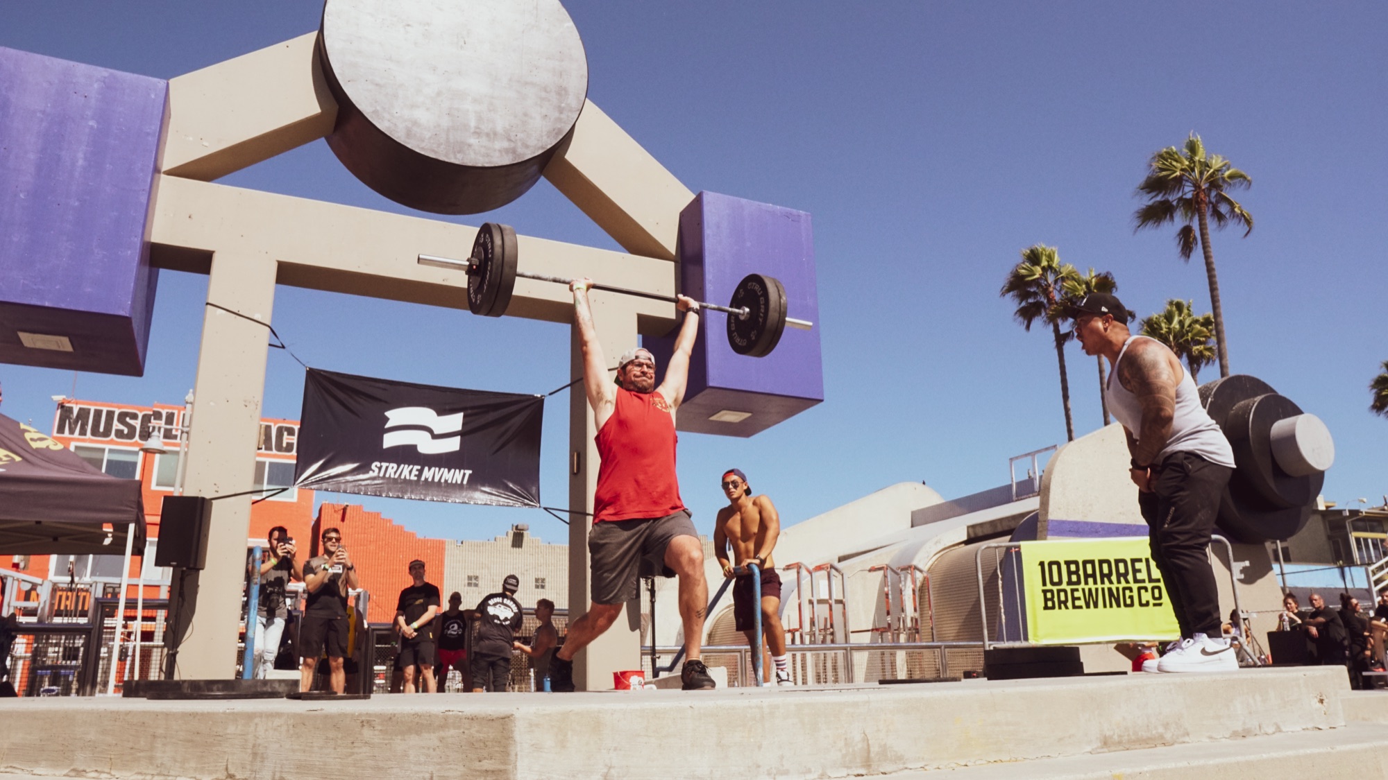 A lifter in a red shirt mid-jerk on the Muscle Beach platform, surrounded by spectators.